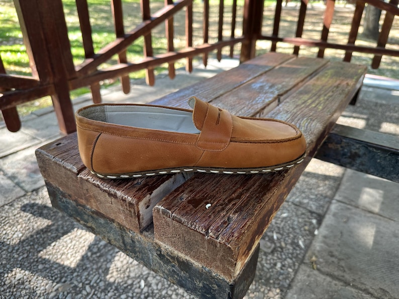 K&ouml;nnte beinhalten: Ein einzelner brauner Leder-Loafer mit Penny-Riemen-Detail, der auf einer verwitterten Holzbank liegt. Der Schuh hat eine wei&szlig; gen&auml;hte Sohle und ein klassisches Design. Der Hintergrund zeigt eine Holzkonstruktion und etwas Gr&uuml;n. Der Schuh ist etwa 25 cm lang.