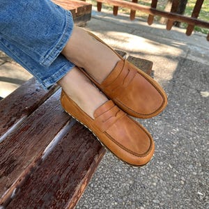 K&ouml;nnte beinhalten: Ein Paar hellbraune Leder-Loafer mit klassischem Penny-Loafer-Design. Die Schuhe haben eine runde Spitze und eine gen&auml;hte Sohle. Die Loafer sind auf einer Holzbank abgebildet.