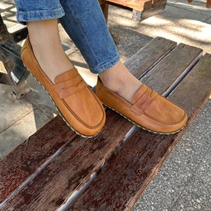 K&ouml;nnte beinhalten: Hellbraune Leder-Loafer mit klassischem Penny-Loafer-Design. Die Schuhe haben eine runde Spitze und eine gen&auml;hte Sohle. Die Loafer sind auf einer Holzbank abgebildet, was auf einen l&auml;ssigen Stil hindeutet.