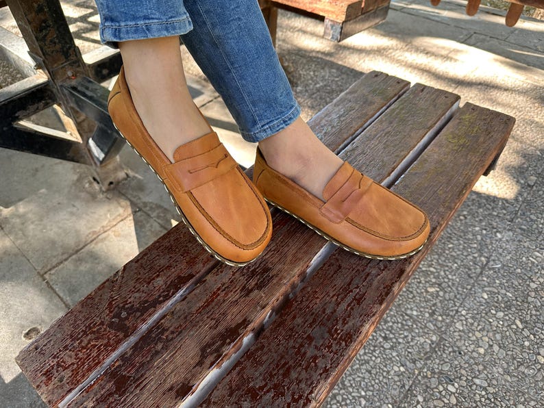 K&ouml;nnte beinhalten: Hellbraune Leder-Loafer mit klassischem Penny-Loafer-Design. Die Schuhe haben eine runde Spitze und eine gen&auml;hte Sohle. Die Loafer sind auf einer Holzbank abgebildet, was auf einen l&auml;ssigen Stil hindeutet.