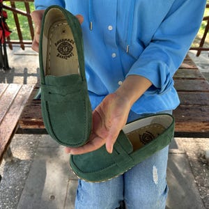 Op de afbeelding: Een paar groene suède loafers. De schoenen hebben een klassiek instapontwerp met een ronde neus en een decoratieve band over de wreef. De binnenkant van de schoen heeft een gedrukt logo met de tekst "HANDMADE BAREFOOT".