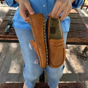 K&ouml;nnte beinhalten: Ein Paar hellbraune Leder-Loafer. Die Schuhe werden von einer Person hochgehalten. Die Loafer haben ein klassisches Design mit runder Spitze, Penny-Riemen und sichtbaren N&auml;hten. Die Sohlen sind dunkler braun.