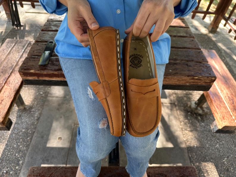 K&ouml;nnte beinhalten: Ein Paar hellbraune Leder-Loafer. Die Schuhe werden von einer Person hochgehalten. Die Loafer haben ein klassisches Design mit runder Spitze, Penny-Riemen und sichtbaren N&auml;hten. Die Sohlen sind dunkler braun.