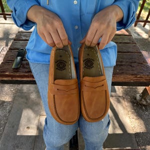 K&ouml;nnte beinhalten: Ein Paar hellbraune Leder-Loafer. Die Schuhe haben ein klassisches Design mit runder Spitze und einem dekorativen Riemen. Die Sohlen sind dunkelbraun. Die Schuhe werden von einer Person gehalten.