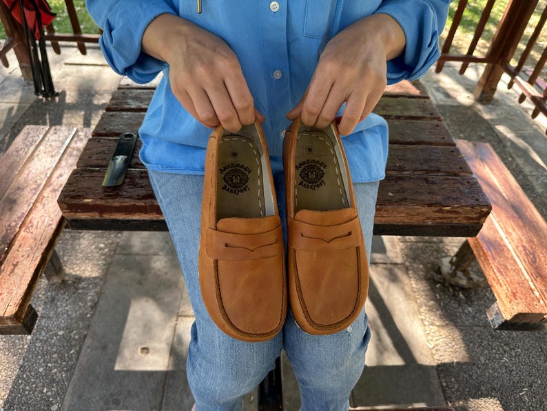 K&ouml;nnte beinhalten: Ein Paar hellbraune Leder-Loafer. Die Schuhe haben ein klassisches Design mit runder Spitze und einem dekorativen Riemen. Die Sohlen sind dunkelbraun. Die Schuhe werden von einer Person gehalten.