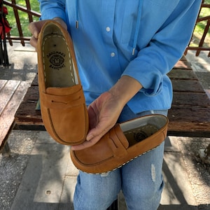 K&ouml;nnte beinhalten: Ein Paar hellbraune Leder-Loafer mit klassischem Design. Die Schuhe haben eine runde Spitze, einen Riemen &uuml;ber der Oberseite und eine gen&auml;hte Sohle. Das Innere des Schuhs hat ein gedrucktes Logo.