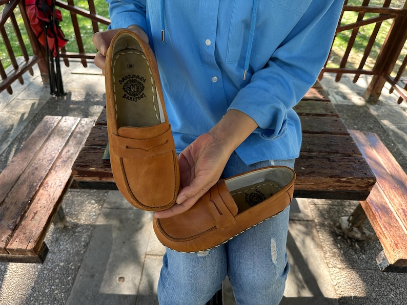 K&ouml;nnte beinhalten: Ein Paar hellbraune Leder-Loafer mit klassischem Design. Die Schuhe haben eine runde Spitze, einen Riemen &uuml;ber der Oberseite und eine gen&auml;hte Sohle. Das Innere des Schuhs hat ein gedrucktes Logo.