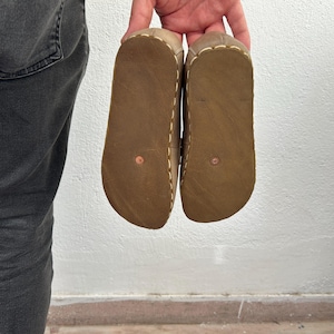 K&ouml;nnte beinhalten: Ein Paar braune Ledersandalen, Sohlen nach oben, vor einer wei&szlig;en Wand gehalten. Die Sandalen haben ein hellbraunes Obermaterial mit sichtbaren N&auml;hten und eine dunkelbraune Sohle. Die Sohlen haben eine kupferfarbene Niete.