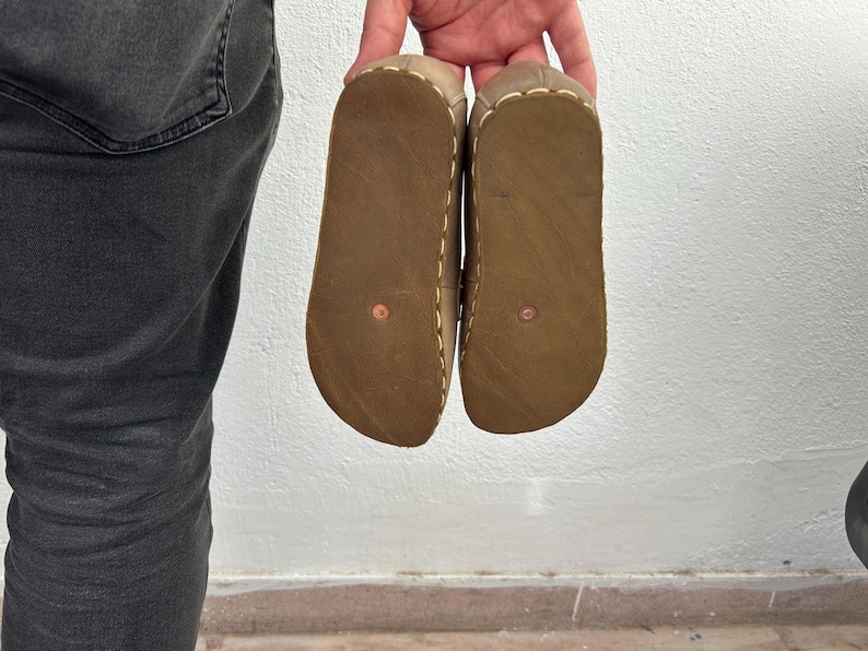 K&ouml;nnte beinhalten: Ein Paar braune Ledersandalen, Sohlen nach oben, vor einer wei&szlig;en Wand gehalten. Die Sandalen haben ein hellbraunes Obermaterial mit sichtbaren N&auml;hten und eine dunkelbraune Sohle. Die Sohlen haben eine kupferfarbene Niete.