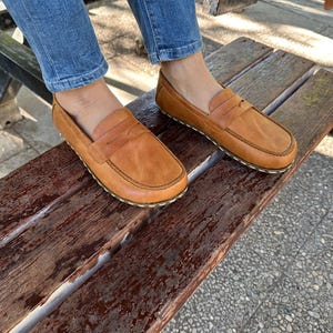 K&ouml;nnte beinhalten: Hellbraune Leder-Loafer mit klassischem Penny-Loafer-Design. Die Schuhe haben eine runde Spitze und eine gen&auml;hte Sohle. Die Loafer werden auf einer Holzbank pr&auml;sentiert und unterstreichen ihren l&auml;ssigen Stil.