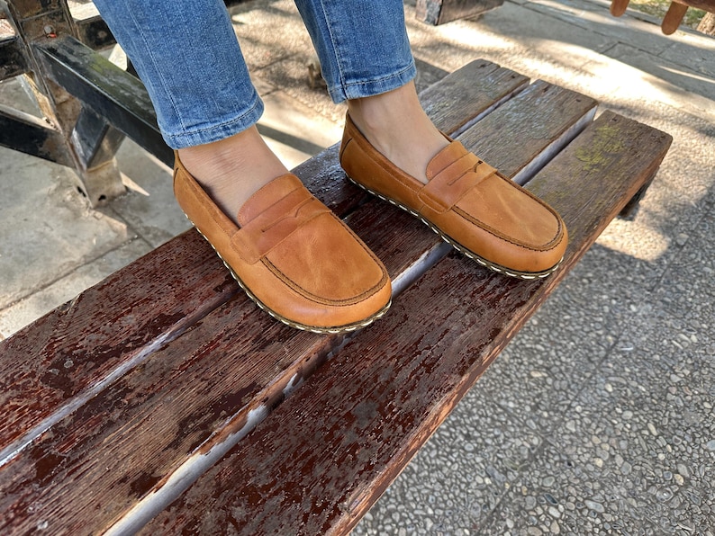 K&ouml;nnte beinhalten: Hellbraune Leder-Loafer mit klassischem Penny-Loafer-Design. Die Schuhe haben eine runde Spitze und eine gen&auml;hte Sohle. Die Loafer werden auf einer Holzbank pr&auml;sentiert und unterstreichen ihren l&auml;ssigen Stil.