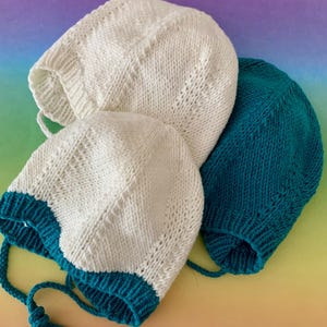 Puede incluir: Tres gorros de bebé tejidos en crema y verde azulado. Dos gorros son crema con ribetes y lazos verde azulado, y el tercero es verde azulado. Los gorros están hechos de un hilo suave y texturizado, sobre un fondo colorido.