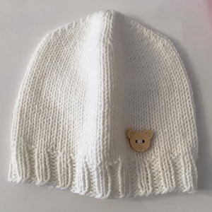 Può includere: Cappellino per neonato in maglia bianca con un piccolo bottone di legno a forma di orso.