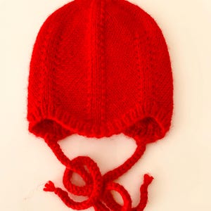 Puede incluir: Un gorro rojo brillante tejido a mano, con orejeras y lazos largos. El gorro está hecho de un hilo suave y texturizado. Los lazos están anudados. Un accesorio de invierno para protegerse del frío.