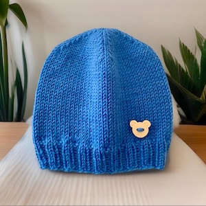 Op de afbeelding: Een blauwe gebreide beanie met een kleine houten berenapplicatie.