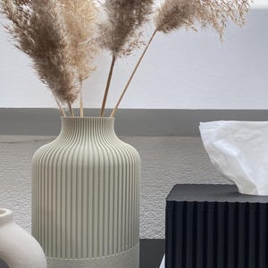 Könnte beinhalten: Dekorative Anordnung mit einer hohen, geriffelten, hellgrünen Vase mit getrocknetem Pampasgras. Eine schwarze Taschentuchbox und ein Stapel Bücher sind ebenfalls zu sehen, was eine minimalistische Ästhetik schafft. Die Vase ist etwa 30 cm hoch.