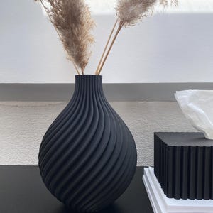Könnte beinhalten: Eine schwarze Vase mit Spiraldesign hält getrocknetes Pampasgras. Die Vase hat eine strukturierte Oberfläche und steht auf einer schwarzen Oberfläche. Eine schwarze Taschentuchbox und weiße Bücher sind ebenfalls sichtbar.