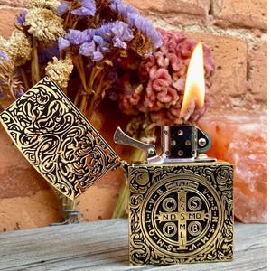 May include: A gold-colored metal lighter with an open lid, displaying intricate engraved patterns. The lighter is lit, with a flame visible. The design includes a cross and text. Dried flowers in purple, pink, and beige are in the background.