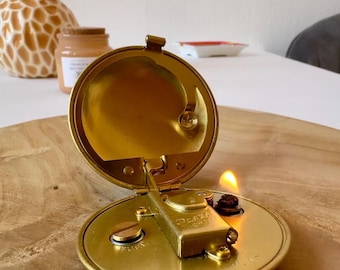 May include: A vintage, open brass lighter with a flame. The lighter is round with a hinged lid and a small, rectangular ignition mechanism. The lighter is on a wooden surface. A vase with flowers is in the background.