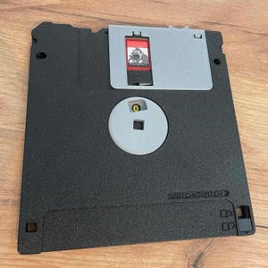 Peut inclure: Un lecteur de disquette noir avec un couvercle coulissant gris et un moyeu circulaire. Le disque comporte une étiquette rouge avec un personnage de dessin animé et le texte "SWITCHISWITCH".