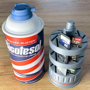 May include: A gray plastic storage container with three rotating tiers for holding memory cards. The container is shaped like a can and has a red and white striped label that reads "BOWSER BUSTER® Gotesol RICH MUSHROOM CREAM ORIGINAL NET QUANTITY 18 CAR".