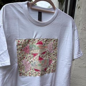 May include: White t-shirt with a floral patch showcasing two fish appliqués. The fish are detailed with pink, green, and cream colours. The shirt is suspended on a wooden hanger, set against a neutral backdrop.