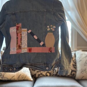 May include: A light blue denim jacket with patches of fabric sewn on the back. The patches depict a bookshelf with books and a vase of flowers. The jacket is hanging on a hanger.