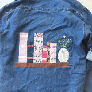 May include: A denim shirt with an embroidered design of a bookshelf with colorful fabric books and a vase with flowers. The books are various patterns and colors, including pink, floral, and blue.