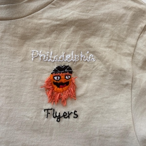 Op de afbeelding: Beige t-shirt met geborduurd ontwerp. Het ontwerp toont het woord "Philadelphia" boven een cartoonmascotte met oranje vacht, een zwart gezicht en een zwarte hoed. Onder de mascotte staat het woord "Flyers" in zwart.