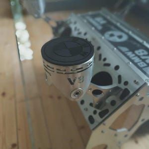 Könnte beinhalten: Einzigartiger Tisch mit einem BMW V8-Motorblock als Basis und einer Glasplatte. Ein Kolben ist sichtbar, mit einem schwarzen Deckel mit dem BMW-Logo und dem Text "V8". Der Motorblock ist silberfarben. Ein originelles Möbelstück.