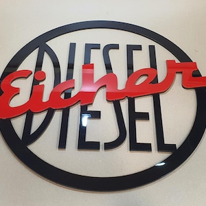 Könnte beinhalten: Ein schwarz-rotes Schild mit den Worten "Eicher Diesel" in einem kreisförmigen Design. Das Wort "Eicher" ist in roter Schrift, und "DIESEL" ist in schwarzen Blockbuchstaben. Das Schild hat einen schwarzen Rand.