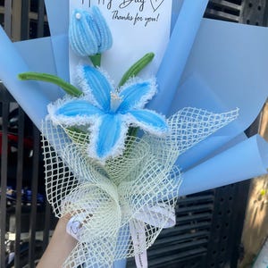 Puede incluir: Un ramo de flores azul y blanco envuelto en papel de seda azul y una red blanca. El ramo tiene una tarjeta que dice "Happy Day" con un corazón y "¡Gracias por ti!"
