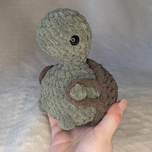 May include: A handmade, plush turtle in shades of green and brown. The turtle has a textured, crocheted appearance, with a rounded body and a small head. The eyes are black. The turtle is held in a hand.