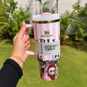 May include: A pink and white striped Stanley Quencher H2.0 tumbler with a handle. The tumbler features a cartoon graphic at the bottom and the Stanley logo. It holds 40 oz (1.18L).