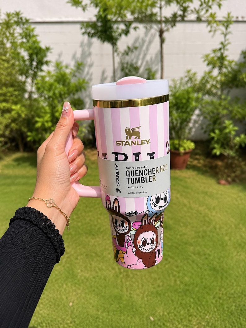 May include: A pink and white striped Stanley Quencher H2.0 tumbler with a handle. The tumbler features a cartoon graphic at the bottom and the Stanley logo. It holds 40 oz (1.18L).