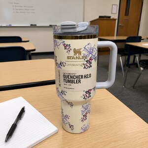 May include: A cream-coloured Stanley Quencher H2.0 tumbler with a floral pattern of purple bows and pink flowers. The tumbler has a grey handle and lid, and the text "LOVESHACKFANCY" and "QUENCHER H2.0 TUMBLER" are visible.