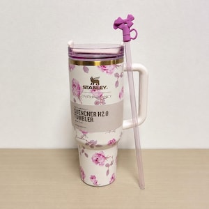 Op de afbeelding: Een witte Stanley Quencher H2.0 tumbler met een bloemenrozenpatroon en een paarse deksel. De tumbler heeft een handvat en een doorzichtige rietje met een paars strikdetail. De tekst "STANLEY LOVESHACKFANCY QUENCHER H2.0 TUMBLER" is zichtbaar.