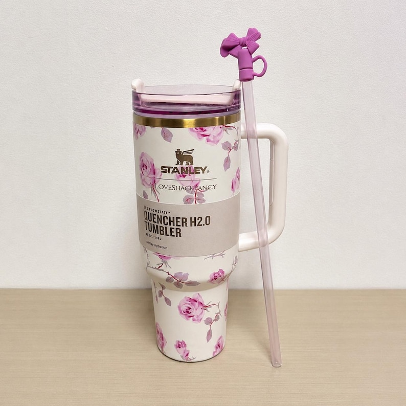 Op de afbeelding: Een witte Stanley Quencher H2.0 tumbler met een bloemenrozenpatroon en een paarse deksel. De tumbler heeft een handvat en een doorzichtige rietje met een paars strikdetail. De tekst "STANLEY LOVESHACKFANCY QUENCHER H2.0 TUMBLER" is zichtbaar.
