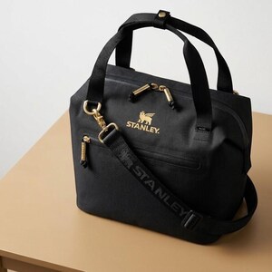 Puede incluir: Bolso tote negro con cremalleras y herrajes dorados. El bolso tiene una correa para el hombro desmontable con el nombre de la marca "STANLEY" impreso. El logotipo de Stanley se muestra en dorado en la parte delantera del bolso.