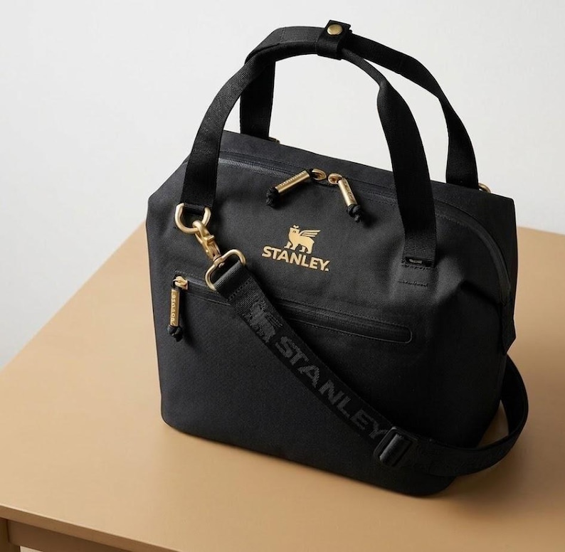 Puede incluir: Bolso tote negro con cremalleras y herrajes dorados. El bolso tiene una correa para el hombro desmontable con el nombre de la marca "STANLEY" impreso. El logotipo de Stanley se muestra en dorado en la parte delantera del bolso.