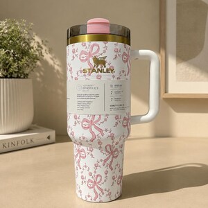May include: A white Stanley Quencher H2.0 tumbler with a pink bow and floral pattern. The tumbler has a pink lid and handle. The Stanley logo is visible. The tumbler is cup holder friendly, spill-resistant, and well-insulated.