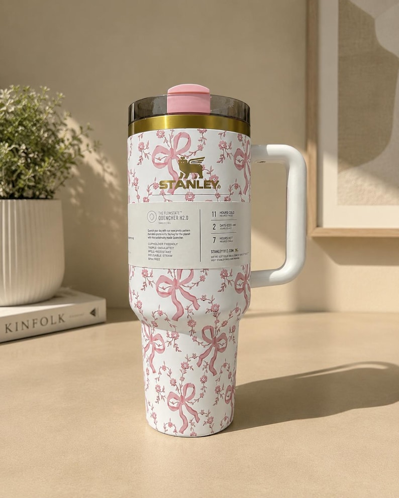 May include: A white Stanley Quencher H2.0 tumbler with a pink bow and floral pattern. The tumbler has a pink lid and handle. The Stanley logo is visible. The tumbler is cup holder friendly, spill-resistant, and well-insulated.