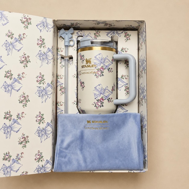 May include: A cream-coloured Stanley LoveshackFancy mug with a floral and bow pattern, a light blue handle, and a matching straw. The mug is presented in a decorative box with a matching floral design and a blue velvet pouch.