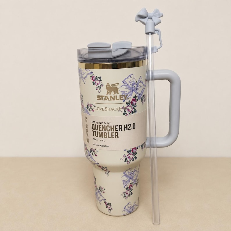 May include: A cream-coloured Stanley Quencher H2.0 tumbler with a floral design featuring pink and purple flowers and bows. The tumbler has a grey handle, lid, and straw with a bow detail. The text "Stanley Loveshack" and "Quencher H2.0 Tumbler" are visible.