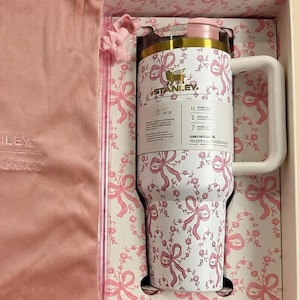 May include: A white Stanley tumbler with a pink floral and bow pattern, presented in a matching gift box with a pink drawstring bag. The tumbler has a white handle and a pink lid. The box and bag are also pink.