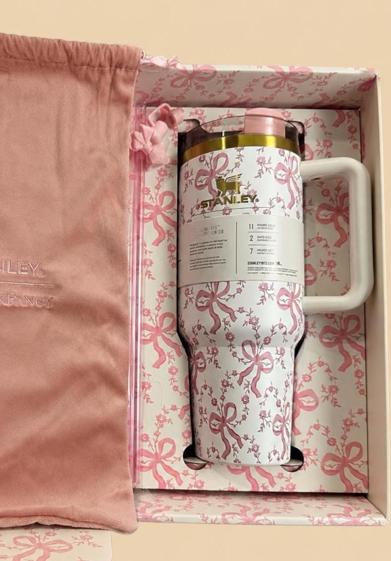 May include: A white Stanley tumbler with a pink floral and bow pattern, presented in a matching gift box with a pink drawstring bag. The tumbler has a white handle and a pink lid. The box and bag are also pink.
