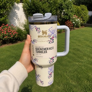 May include: Cream-coloured Stanley Quencher H2.0 tumbler with a floral pattern of pink and purple flowers and bows. The tumbler has a light grey handle and lid. Text on the tumbler reads "Stanley", "LoveShack", and "Quencher H2.0 Tumbler".