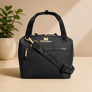 Puede incluir: Bolso tote negro aislado con cremalleras y herrajes dorados. Cuenta con un asa superior y una correa para el hombro desmontable con el nombre de la marca "STANLEY" impreso. El bolso tiene el logotipo "STANLEY" en dorado.