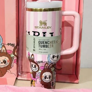 May include: A pink Stanley Quencher H2.0 tumbler with a white lid and handle, presented in a matching box. The tumbler features a striped design and cartoon graphics. The capacity is 40 oz.