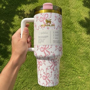May include: A white Stanley Quencher H2.0 Flowstate tumbler with a pink bow and floral pattern. The tumbler has a handle, a pink lid, and gold accents. The Stanley logo is visible. The tumbler is designed to keep drinks cold for 11 hours.